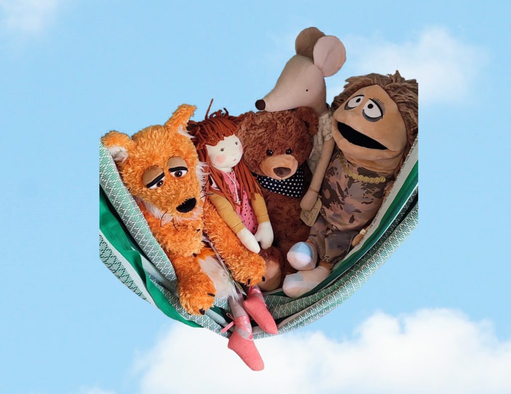 Verschiedene Stofftiere und Stoffpuppen liegen gemeinsam in einer Hängematte vor blauem Himmel, Symbolbild für Geborgenheit und Verbundenheit im Workshop "Spielräume der Seele"