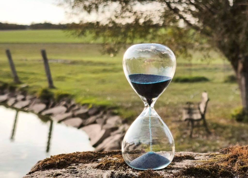 Eine Sanduhr mit dunklem Sand steht im Abendlicht auf einem Stein am Wasser, Symbolbild für die klar strukturierten Schritte und die transparenten Kosten einer Zusammenarbeit
